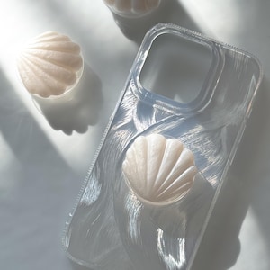 May include: A clear phone case with a seashell design and a matching seashell phone grip. Two additional seashell-shaped objects are also visible. The items are white and appear to be made of plastic or resin.