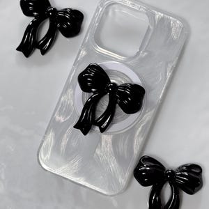 Könnte beinhalten: Eine transparente Handyhülle mit einem schwarzen Schleifendesign. Die Hülle hat eine strukturierte Oberfläche und einen Ringhalter mit einer schwarzen Schleife.