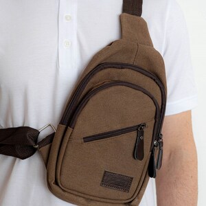 May include: A brown canvas crossbody bag with a zippered pocket and a brown leather patch. The bag has a brown adjustable strap.