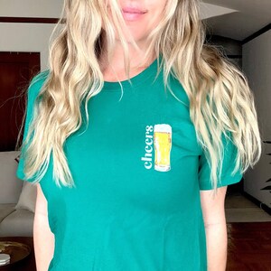 Op de afbeelding: Een groen T-shirt met het woord "cheers" en een afbeelding van een glas bier.