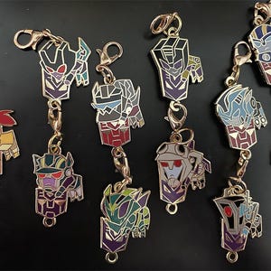 May include: A collection of colorful metal keychains featuring various robot character designs. Each keychain has a gold-colored clasp and chain. The robots are depicted in a variety of colors, including red, purple, green, and blue.