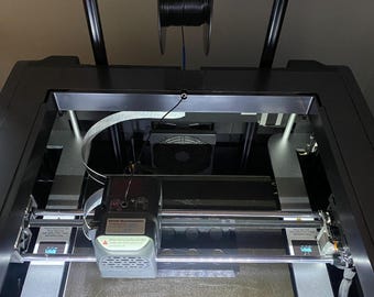 May include: A black 3D printer with a spool of black filament on a holder above the printer bed. The printer bed is lit with a white light.