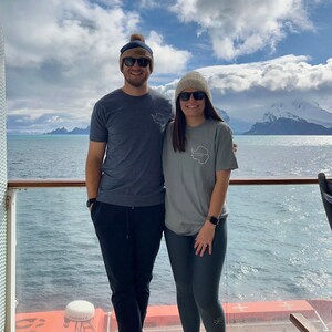 May include: A couple wearing matching grey t-shirts with a white outline of the continent of Antarctica on the front. They are standing on a cruise ship deck with a view of the ocean and mountains in the background.