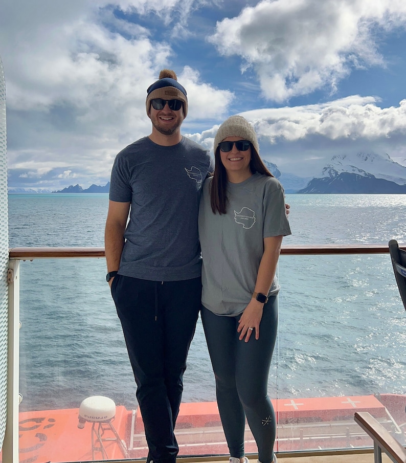 May include: A couple wearing matching grey t-shirts with a white outline of the continent of Antarctica on the front. They are standing on a cruise ship deck with a view of the ocean and mountains in the background.