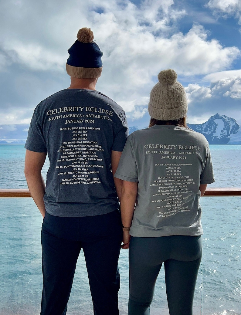 May include: Two people wearing grey t-shirts with the text "Celebrity Eclipse South America - Antarctica January 2024" and a list of dates and locations. The people are standing on a deck overlooking a body of water with mountains in the background.