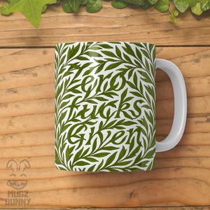 May include: White ceramic mug with a green leaf pattern and the words "Zero Fucks Given" in a stylized font. The mug has a white handle and is set against a wooden background with green foliage.