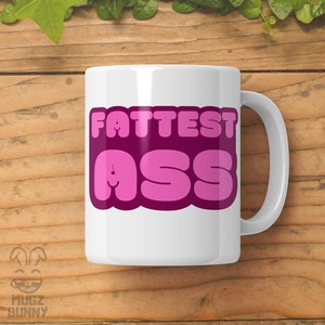 May include: White ceramic mug with a pink and purple graphic that says "FATTEST ASS" in a bubble font.