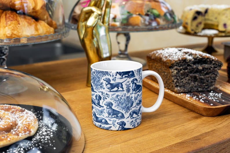 May include: A white ceramic mug featuring a blue and white pattern of dachshunds. The mug is on a wooden table with other items in the background.