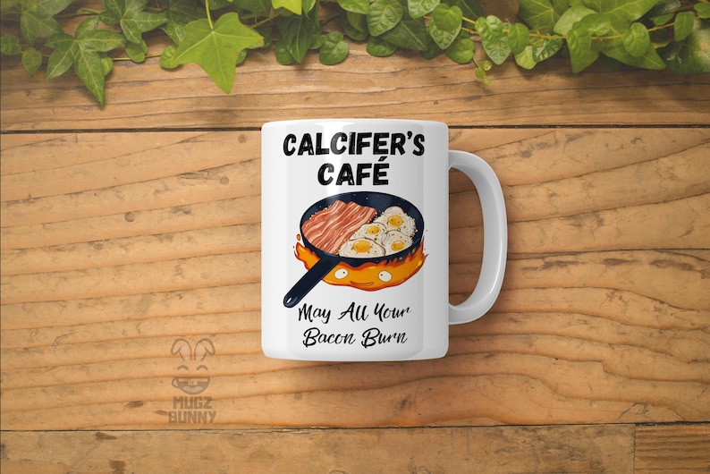 May include: White ceramic mug with a black design featuring a cartoon fire demon holding a frying pan with bacon and eggs. The text reads "Calcifer's Cafe" and "May All Your Bacon Burn".