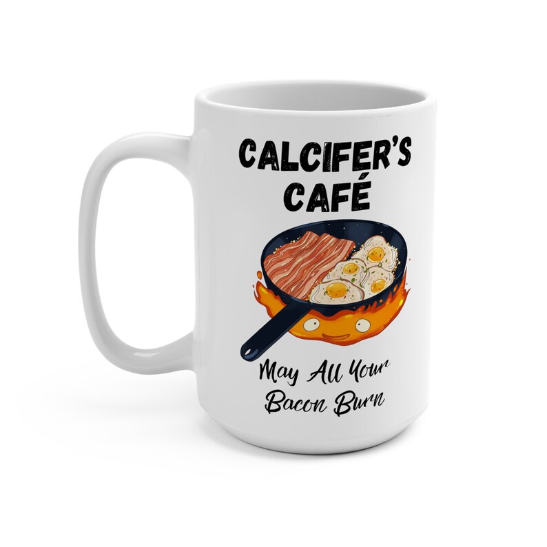 May include: White ceramic mug with a black and white graphic of a cartoon fire demon holding a frying pan with bacon and eggs. The text "Calcifers Cafe" and "May All Your Bacon Burn" is printed on the mug.