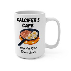 May include: White ceramic mug with a black and white illustration of a frying pan with bacon and eggs on fire. The text "Calcifer's Cafe" and "May All Your Bacon Burn" is printed on the mug.