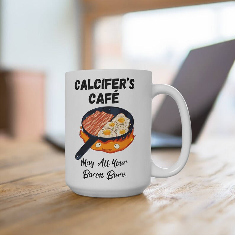 May include: White ceramic mug with a black and white illustration of a cartoon fire demon holding a frying pan with bacon and eggs. The text "Calcifer's Cafe" and "May All Your Bacon Burn" is printed on the mug.