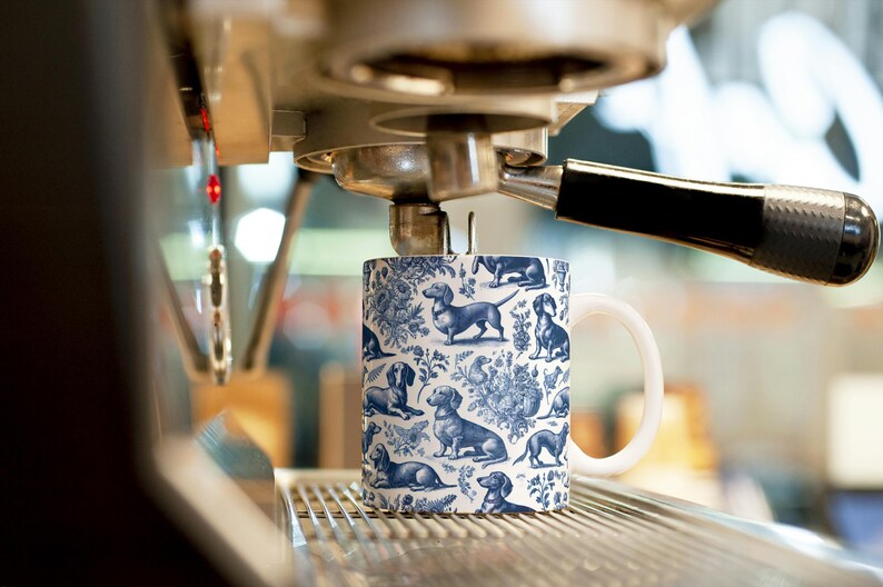 May include: A white ceramic mug with a blue and white pattern of dachshunds. The mug is sitting on a metal rack under a coffee machine.