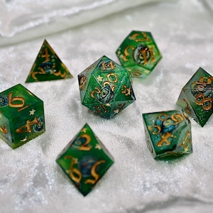 May include: Set of seven green and gold resin dice for tabletop games. Each die features gold numbering and intricate designs within the translucent green resin. The dice are arranged on a white, textured surface, showcasing their multifaceted shapes.