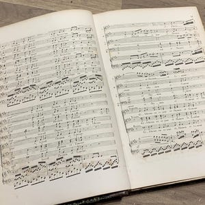 Może przedstawiać: Otwarta książka z wydrukowaną notacją muzyczną na stronach. Książka jest oprawiona w brązową skórę i ma zużyty, staroświecki wygląd.