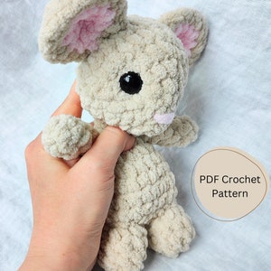 Könnte beinhalten: Ein gehäkeltes Plüschtier-Kaninchen in Beige mit rosa Ohren und einem schwarzen Auge. Das Kaninchen wird von einer Hand gehalten. Der Text "PDF Crochet Pattern" ist im Bild sichtbar.