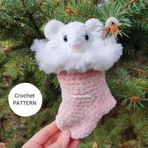 Op de afbeelding: Een roze gehaakte kerstsok met een wit, pluche muizenhoofd erop. De sok is versierd met een witte bontkraag. De sok hangt aan een groene kerstboom.