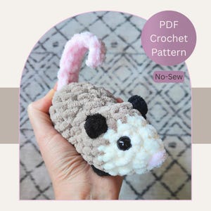 May include: A crocheted opossum toy in shades of gray, white, and pink. The plush animal has black ears and a pink tail. The image includes text that reads "PDF Crochet Pattern" and "No-Sew".