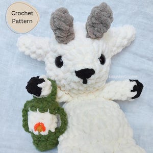 Può includere: Peluche di capra bianca all'uncinetto con occhi e naso neri, che tiene una lanterna verde all'uncinetto con una fiamma arancione all'interno. Il giocattolo ha corna grigie all'uncinetto. Il testo "LolliMayCrochet" è visibile sullo sfondo.
