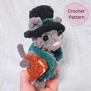 May include: A gray crocheted cat wearing a black top hat, round glasses, and a teal scarf. The cat is holding a small orange crocheted bag.