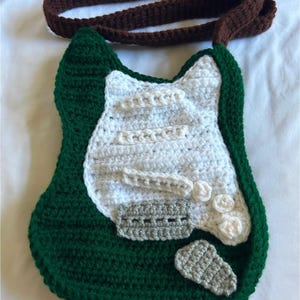 Puede incluir: Una bolsa de ganchillo en forma de guitarra de color verde, blanco y gris con una correa de ganchillo marrón.