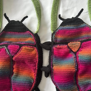 May include: Two handmade crochet bags shaped like beetles. Each bag features a black head with antennae, a triangular orange accent, and a body made of multicolored yarn in shades of pink, orange, purple, and blue. Green crochet straps are attached.