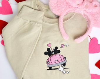 May include: Tan sweatshirt with an embroidered design of two cartoon mice in a pink car, with hearts and flowers. A pair of pink, fluffy mouse ears are next to the sweatshirt. Red and pink heart garland.