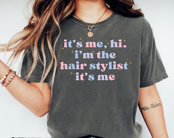 May include: A dark gray t-shirt with the text "it's me, hi. i'm the hair stylist it's me" in pink and blue lettering. The shirt has a distressed look and is worn by a person with long blonde hair.