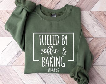 May include: A military green sweatshirt with the text "Fueled by coffee & baking" in a white rectangular frame. The word "#BAKER" is below the text. The sweatshirt is displayed on a wooden surface.