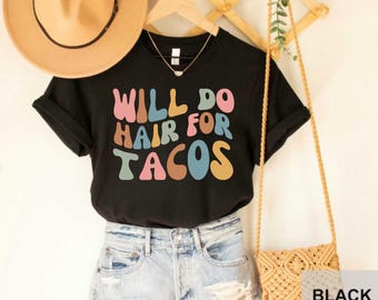 May include: Black t-shirt with the text "Will do hair for tacos" in colorful, retro-style lettering.