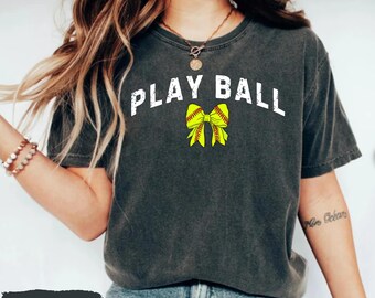 May include: Dark grey heather t-shirt with the text "PLAY BALL" in white block letters. A yellow softball bow graphic is centered below the text. The shirt is a casual style.