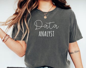 May include: A gray t-shirt with the text "Data Analyst" in white lettering. The shirt is worn by a person with long brown hair. The text "Color: Pepper" is visible at the bottom of the image.