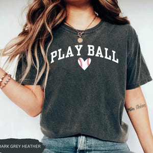 Peut inclure: Un t-shirt en gris chin&eacute; fonc&eacute; avec le texte "PLAY BALL" en lettres blanches et un graphique de c&oelig;ur de baseball rouge.