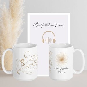 Könnte beinhalten: Zwei weiße Keramikbecher mit goldenen botanischen Designs und den Worten "Manifestation Power". Ein gerahmter Druck hinter den Bechern zeigt ebenfalls die Worte "Manifestation Power" und eine goldene Kopfhörergrafik. Der Hintergrund umfasst Pampasgras.