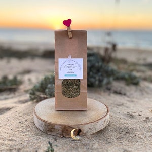 Puede incluir: Una bolsa de papel marrón con una etiqueta blanca que dice "Luftpflanzen Tee" y un pequeño clip en forma de corazón. La bolsa está sentada sobre una rodaja redonda de madera en una playa de arena.