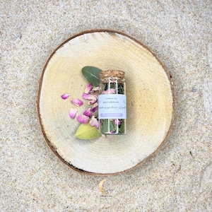 Könnte beinhalten: Eine kleine Glasflasche gefüllt mit getrockneten Kräutern und Blumen, verschlossen mit einem Korken, liegt auf einer Holzscheibe. Die Flasche hat ein Etikett mit Text. Rosa Rosenblätter und grüne Blätter sind um die Flasche auf dem Holz verstreut.