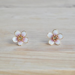 Op de afbeelding: Een paar bloemenknopjes. Elke oorbel heeft een bloemontwerp met vijf bloemblaadjes met witte en roze emaille bloemblaadjes, een gouden omtrek en een centrale gouden zetting met een kleine, heldere edelsteen. Op een licht houten oppervlak.