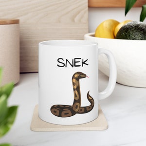 May include: White ceramic mug with the word "SNEK" in black letters and a cartoon snake illustration. The snake is brown and tan with a red tongue. The mug sits on a beige coaster.