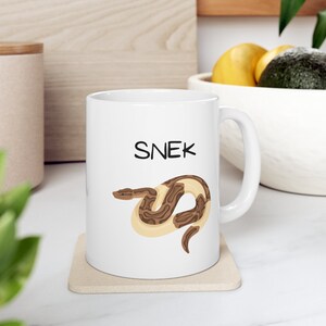 May include: White ceramic mug with a brown and white snake illustration and the word "SNEK" in black text.