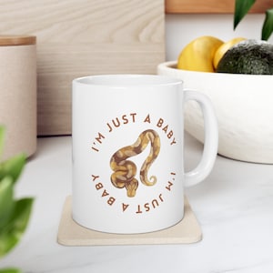 May include: White ceramic mug with a brown snake illustration and the text "I'm just a baby I'm just a baby" in a circle around the snake.