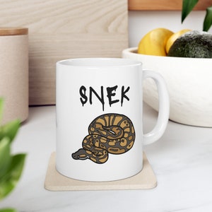 May include: White ceramic coffee mug with a black snake illustration and the word "SNEK" in black lettering.