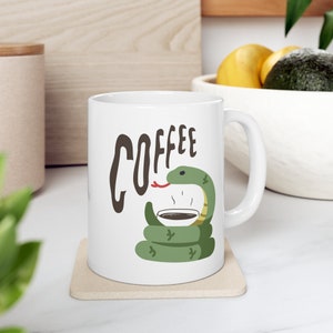 May include: White ceramic coffee mug with a green snake wrapped around it, drinking coffee from a cup. The mug has the word "COFFEE" written on it in brown letters.