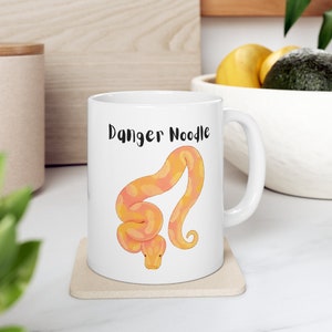 May include: White ceramic mug with a yellow and orange snake illustration and the text "Danger Noodle" printed on the front.