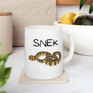 May include: White ceramic coffee mug with a brown snake illustration and the word "SNEK" printed in black.