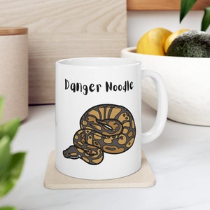 May include: White ceramic mug with a black and brown cartoon snake illustration and the text "Danger Noodle".
