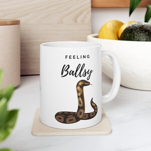 May include: White ceramic mug with a brown and black snake illustration and the text "Feeling Ballsy".
