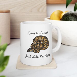 May include: White ceramic coffee mug with a snake illustration. The mug has the text "Spicy & Smooth, Just Like My Coffee" in black script. The snake is brown and tan with a black outline. The mug sits on a beige coaster.