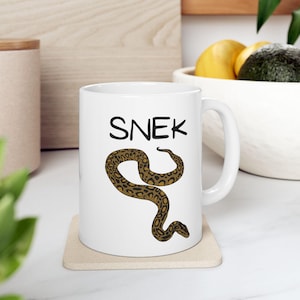 May include: White ceramic mug with a brown snake illustration and the word "SNEK" in black text.