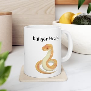 May include: White ceramic mug with a yellow snake illustration and the text "Danger Noodle" printed on the front.