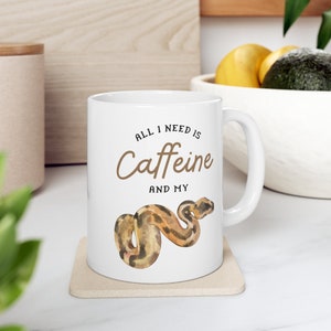May include: White ceramic mug with a brown snake illustration and the text "All I need is Caffeine and my".